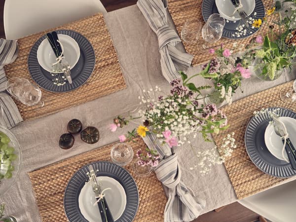 Vue aérienne d'une table dressée avec des assiettes grises, des bols blancs, des fleurs, des verres et des sets de table tissés.