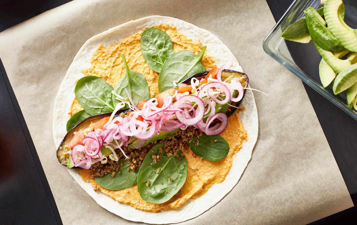 Vue aérienne d’une tortilla posée sur du papier de cuisson et garnie entre autres avec du houmous, des épinards, une aubergine rôtie et des oignions pour faire un burrito. 