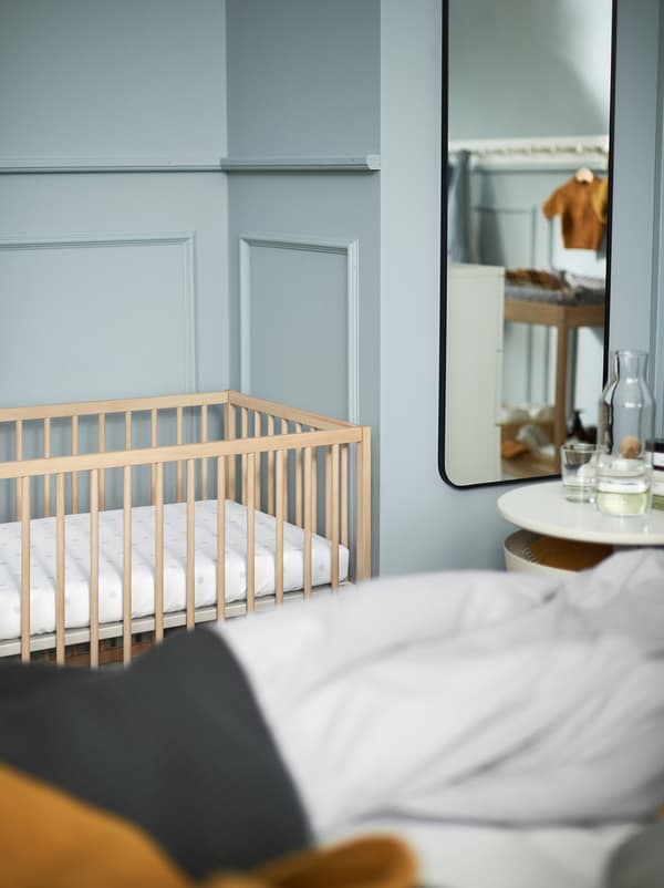 Vue partielle d’une chambre bleu clair avec une table de chevet blanche, un miroir et un lit bébé en bois SNIGLAR.