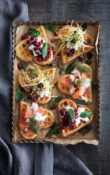 Waffles with various toppings, such as marinated salmon, dried cranberries with a mix of cress, leek and carrots and pomegranate seeds mixed with cottage cheese.