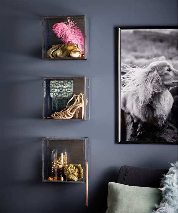 Wall-mounted BARKHYTTAN display boxes feature a large pink feather, gold high heels, and other assorted items.