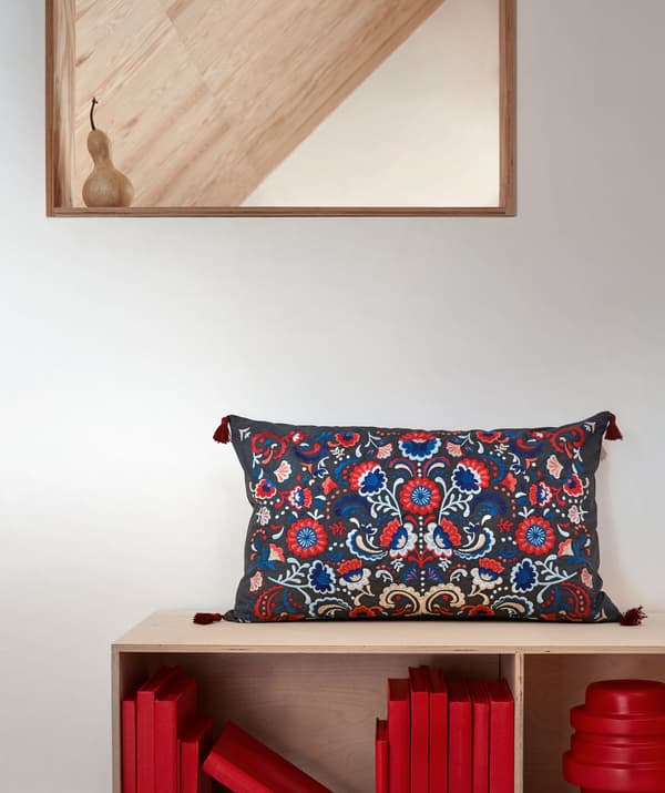 Wall section with mirror and a low bookcase below, with a colourful, embroidered SKOGSKORN cushion on top.