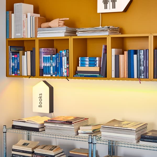 Wall storage with several EKET open cubes in yellow filled with books, and metal storage standing on the floor underneath it.