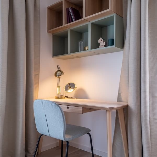 Warm and inviting home workspace with a natural wood desk and a soft blue upholstered chair. The desk is lit by a stylish gold desk lamp and a small round accent lamp, casting a cozy glow. Above the desk are open wall-mounted shelves in a mix of wood and sage green tones, holding decorative items such as books, a candle, a small birdhouse, and a glass bottle. Beige curtains frame the setup, enhancing the calm and modern atmosphere of this compact study nook.