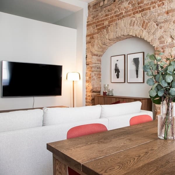 Warm and inviting living-dining area combining modern furniture with rustic architectural elements. A dark wooden dining table with red-upholstered chairs is in the foreground, decorated with a glass vase of eucalyptus branches. Behind it, a light beige sectional sofa faces a wall-mounted flat-screen TV and a modern floor lamp. The exposed brick archway adds texture and character to the room, framing two minimalist black-and-white art prints. Wooden cabinetry and soft lighting contribute to the cozy, stylish ambiance.