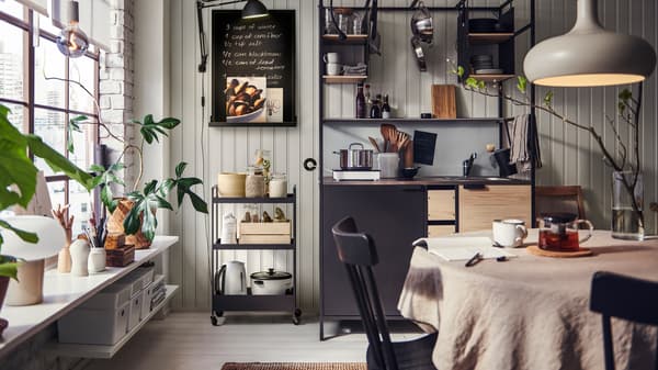 Warm kitchen and dining area with open shelving, wooden table, hanging lights, plants, appliances and a cozy, lived-in feel.