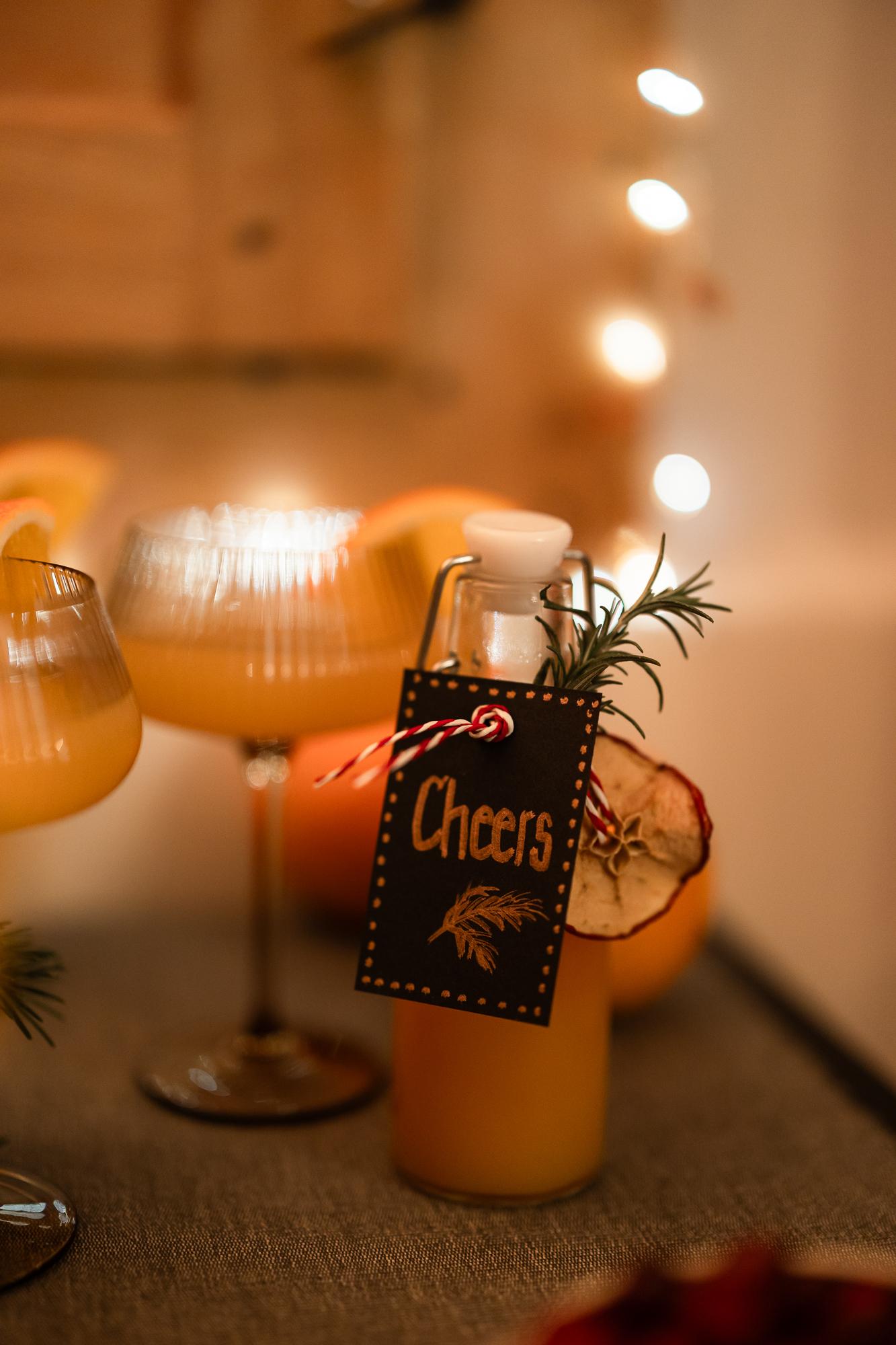 Warme, festliche Szene mit orangefarbenen Getränken in eleganten Gläsern. Eine Flasche mit einem „Cheers“-Etikett, garniert mit Rosmarin und getrockneter Orange, sanfte Beleuchtung im Hintergrund.