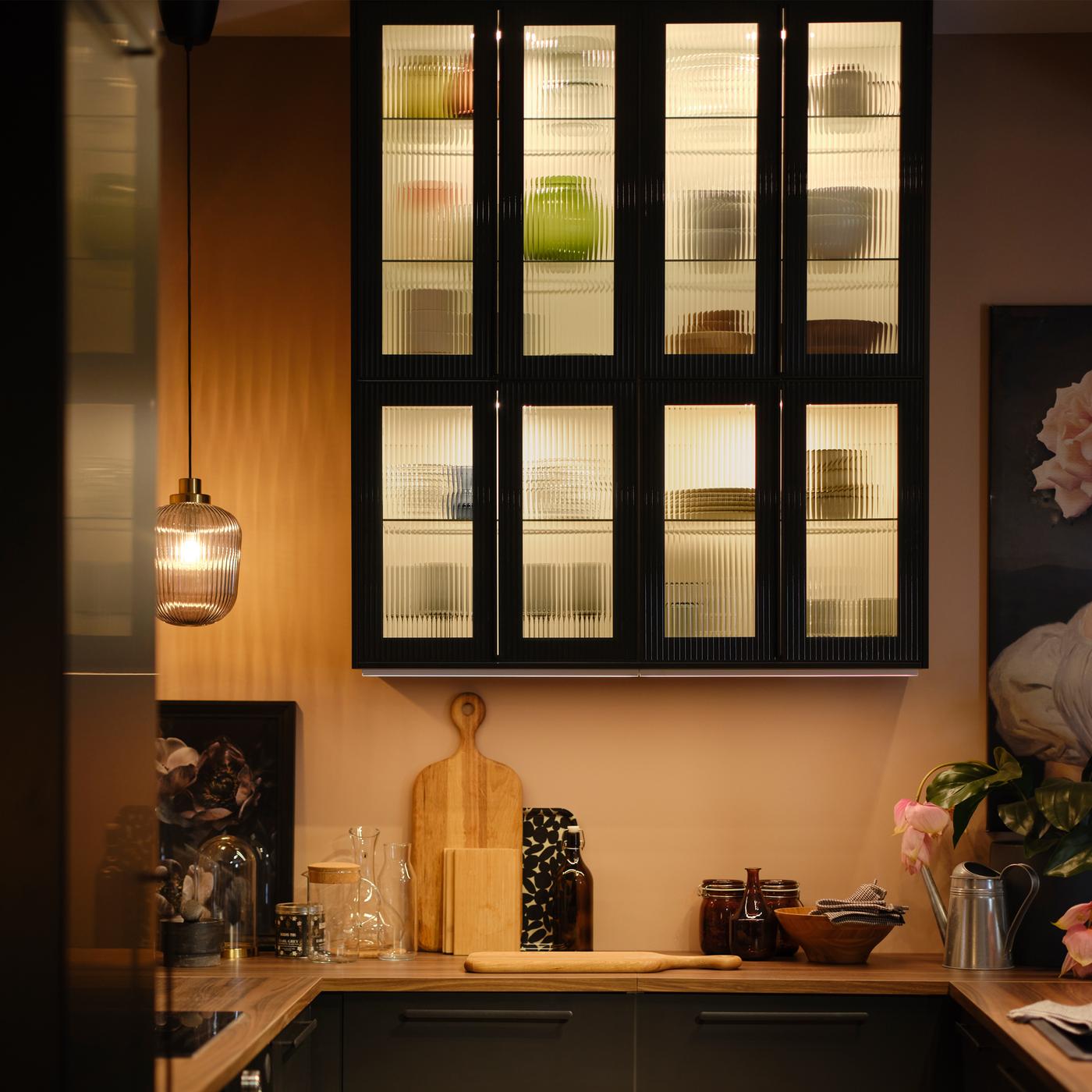 White METOD wall cabinets with shelves and anthracite HEJSTA reeded glass doors with various items and lighting inside.