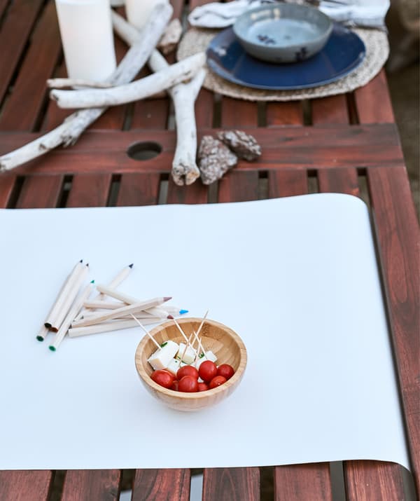 White paper and coloured pencils and a small bowl of snacks on a wooden table, next to driftwood and a place setting.