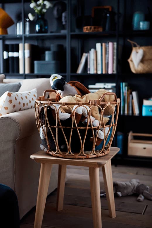 Wicker basket, standing on a stool in a living room, filled to the brim with different soft toy animals.