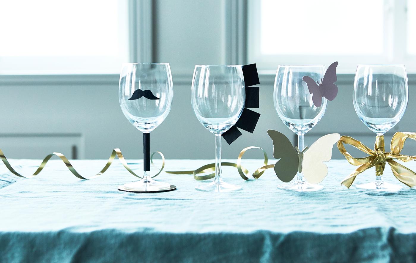 Wine glasses decorated with various home-made paper decorations on a table with a blue tablecloth.