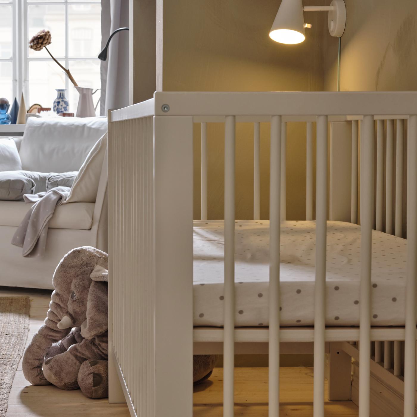 Wit GULLIVER babybed met een pluchen olifant aan één uiteinde en een witte fauteuil erachter. De wandlamp ernaast brandt.