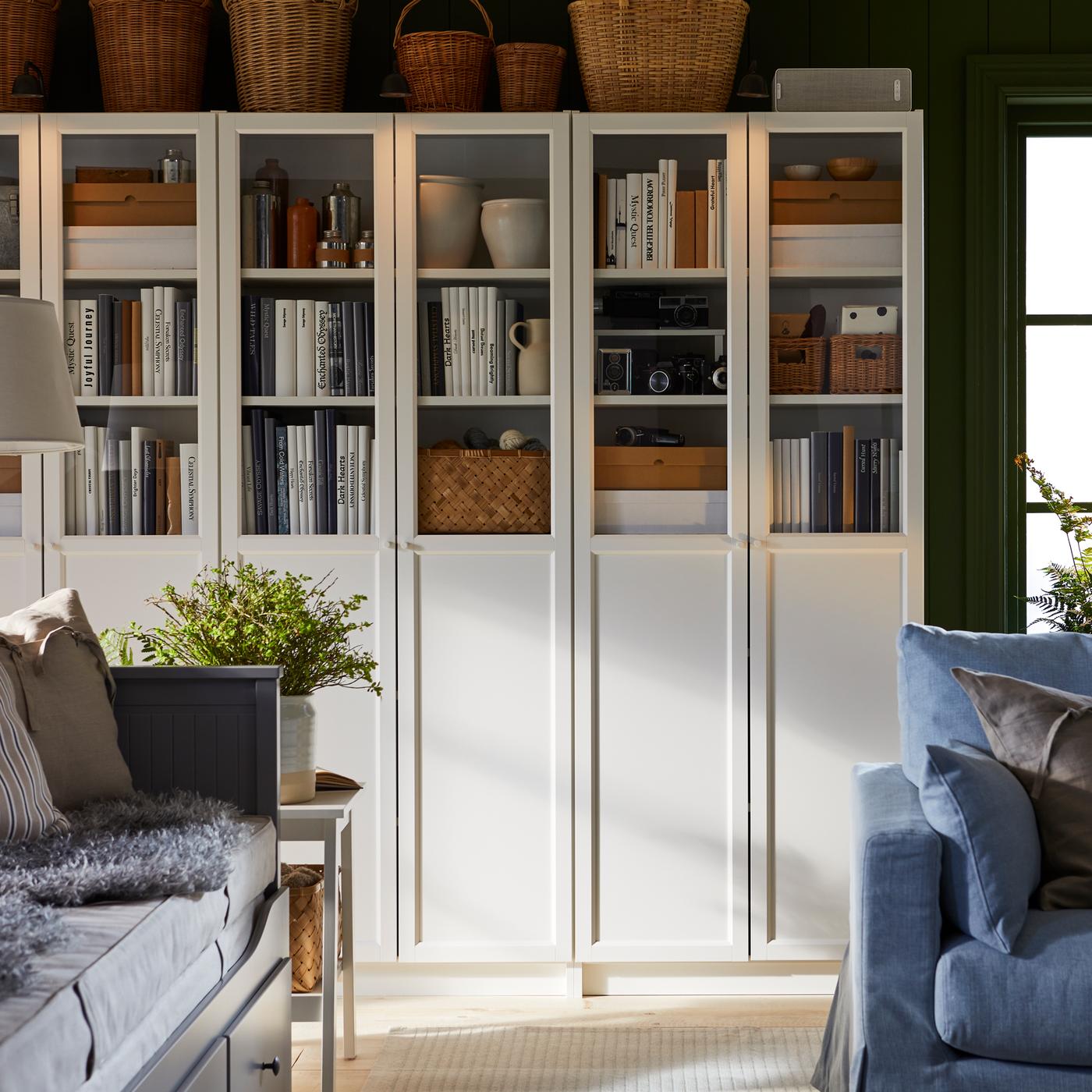 Witte BILLY boekenkasten met paneel-/vitrinedeuren staan vol boeken, dozen en snuisterijen, naast een raam in een woonkamer.