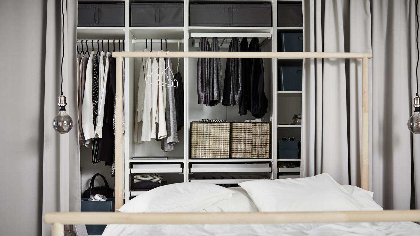 Witte PAX kledingkastcombinatie met verschillende vakken waarin verschillende kledingstukken liggen, achter een bed met een wit dekbed.