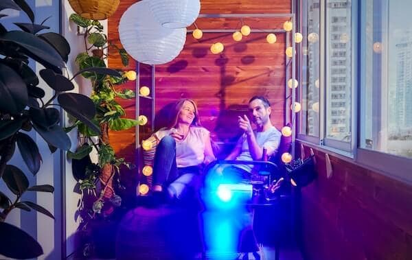 Woman and a man sitting on a balcony in a SVANÖ arbor bench with a running film projector on a side table in front of them.