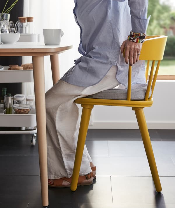 Woman in a semi-seated position using her hands to gain support from the sturdy back of a yellow OMTÄNKSAM chair.