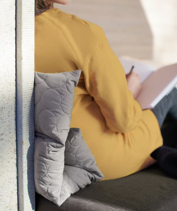 Woman leaning back, sitting with a folded, grey OMTÄNKSAM multi-cushion.
