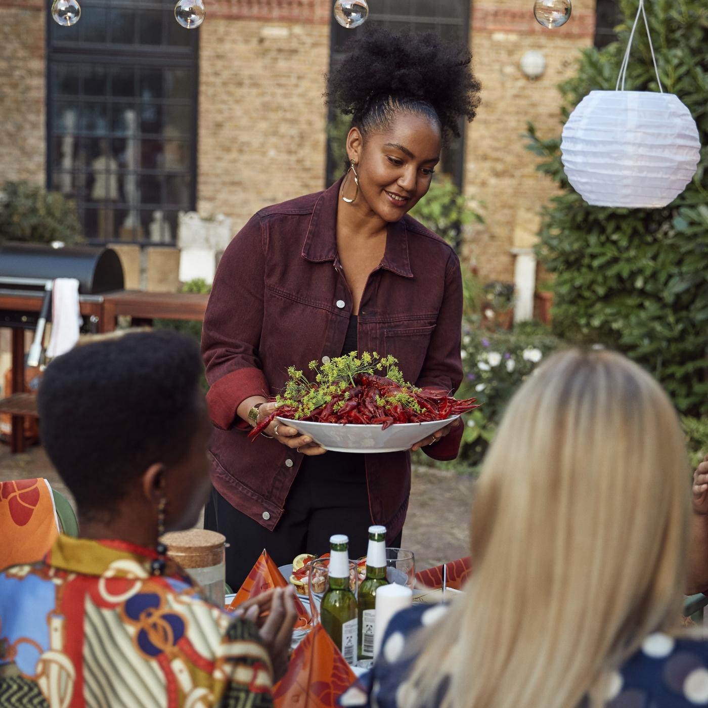 Woman serving crayfish in a white bowl for friends sitting around a table propped with sparkling drinks and crayfish-themed napkins.