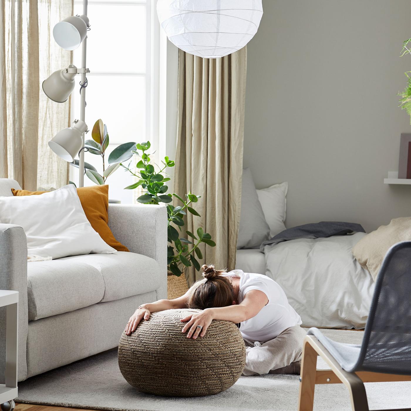 Woman sitting on a living room rug, legs folded under her and stooped over, her outstretched arms resting on a pouffe.