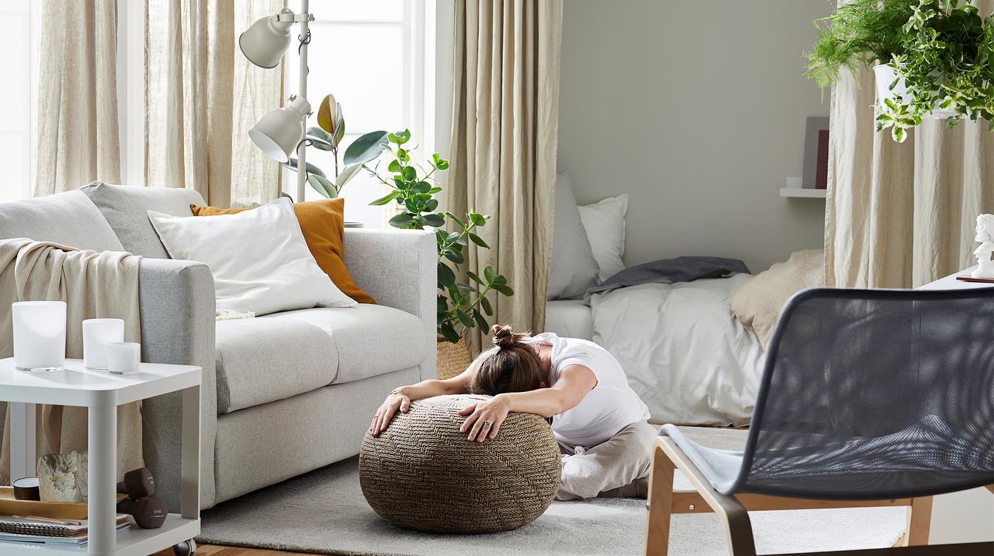 Woman sitting on a living room rug, legs folded under her and stooped over, her outstretched arms resting on a pouffe.
