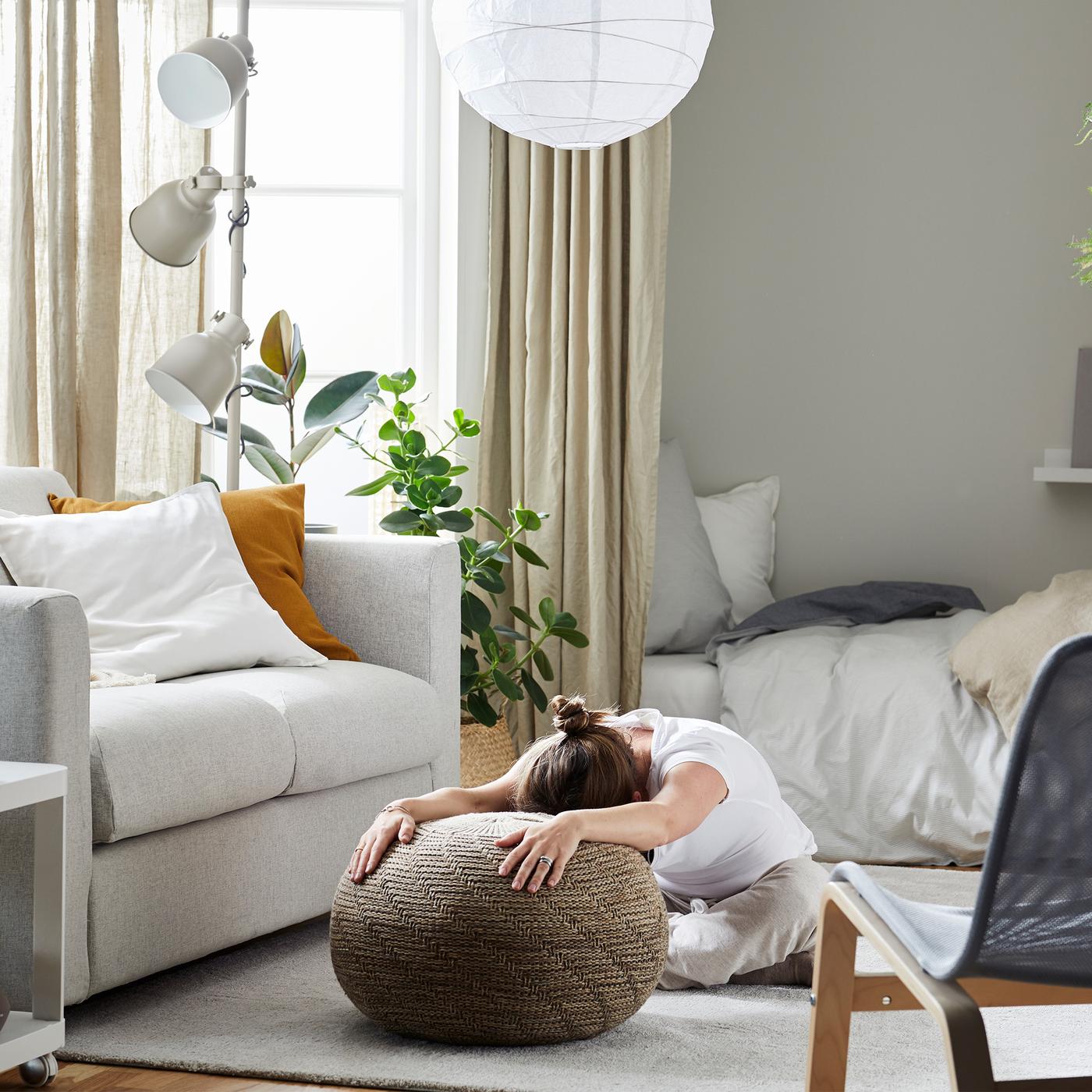 Woman sitting on a living room rug, legs folded under her and stooped over, her outstretched arms resting on a pouffe.