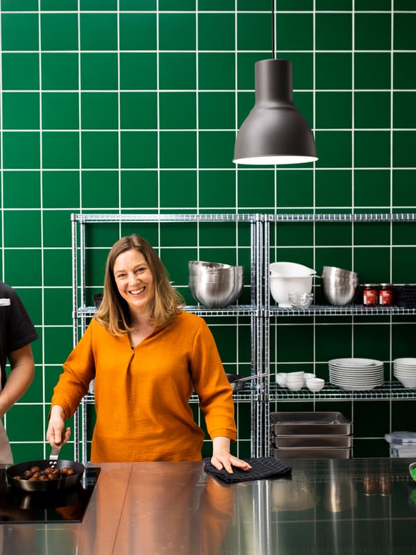 Woman standing in a kitchen styled with stainless steel and green wall tiles, holding a frying pan of plant-based meatballs.