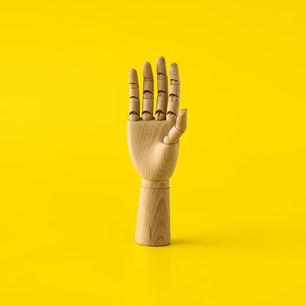 Wooden mannequin hand raised upright with fingers extended, positioned against a bright yellow background.