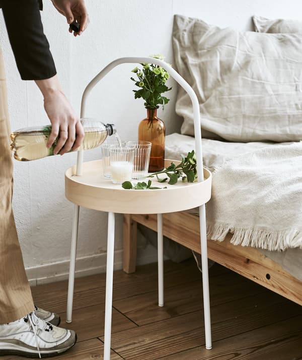 Yvet pours a drink into a glass on a light wood side table with white handle next to a bed.