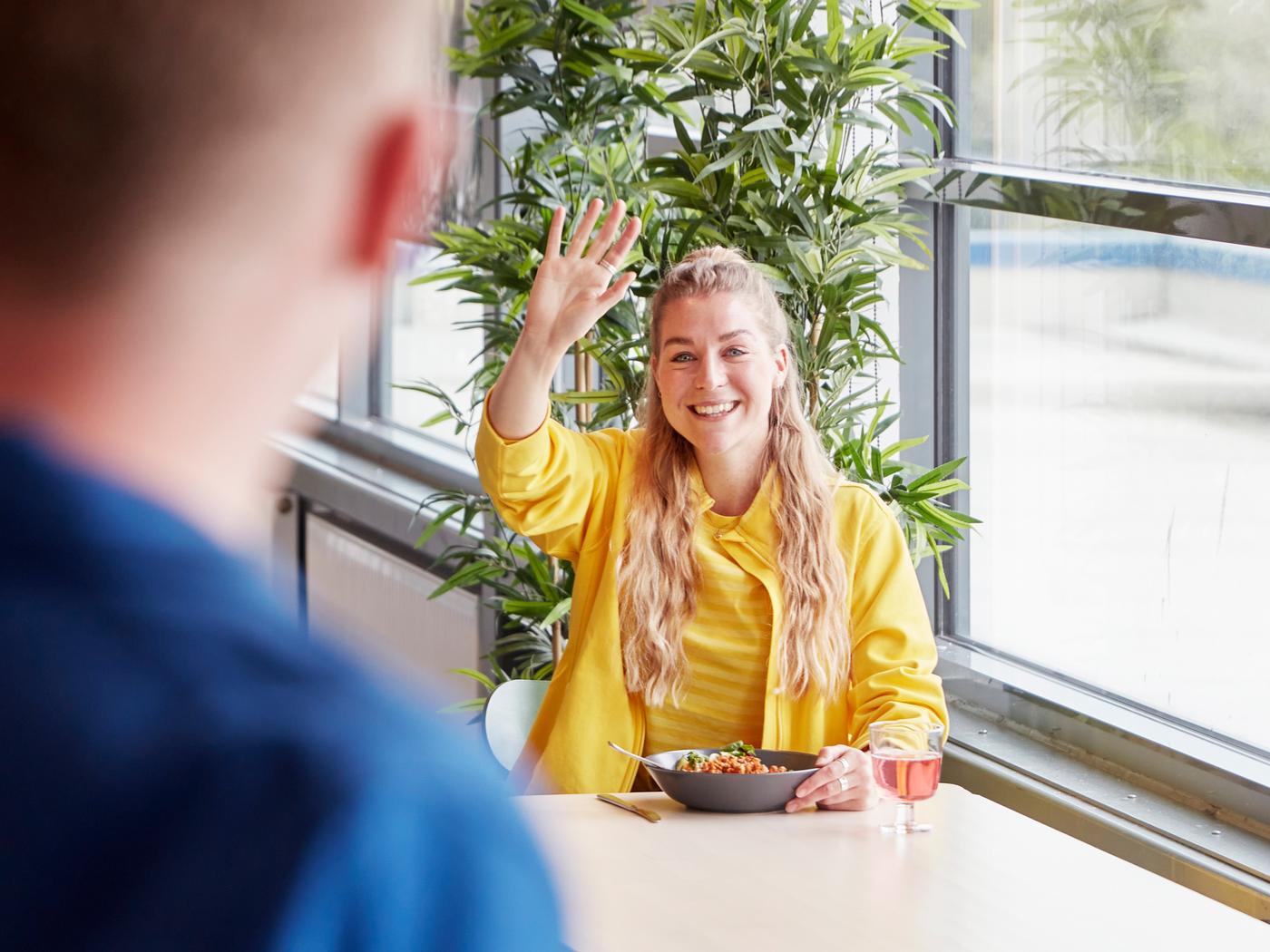 Zaměstnankyně IKEA ve žluté uniformě mává kolegovi v jídelně pro zaměstnance.