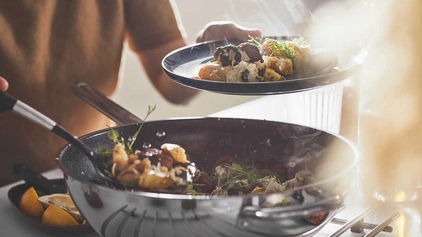 Žena v béžovém svetru nandavá z pánve wok IKEA 365+ na talíř směs kuliček a zeleniny.