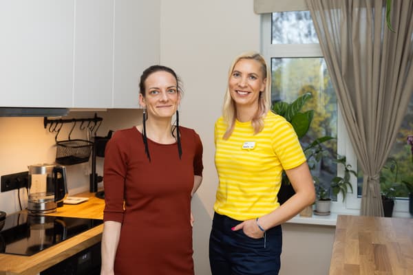 Женщина в красном платье и дизайнер IKEA в жёлтой форме стоят на кухне с белыми фасадами и деревянной столешницей.