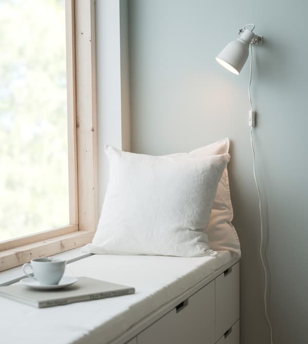 Zitplekje aan het raam van een slaapkamer, gemaakt van witte NORDLI ladekasten, twee witte kussens en een dunne dekmatras