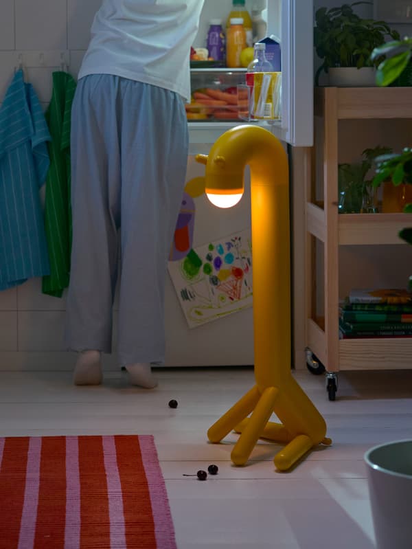 Žlutá stojací lampa ve tvaru žirafy GREJSIMOJS stojí v kuchyni, za ní někdo pátrá v otevřené ledničce.