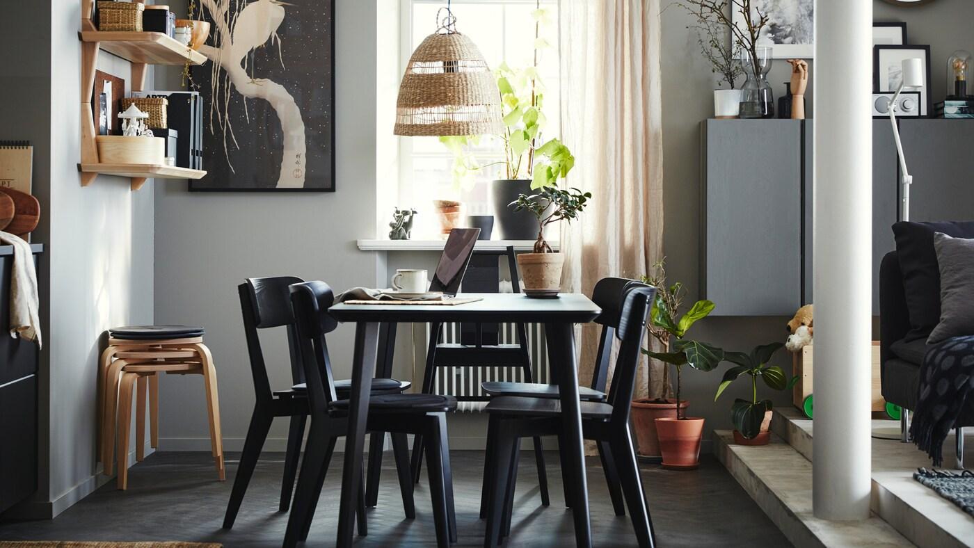 Zona pranzo con tavolo, quattro sedie e un seggiolone, tutto in nero. Scaffali in impiallacciatura di frassino, quattro sgabelli e tende beige.