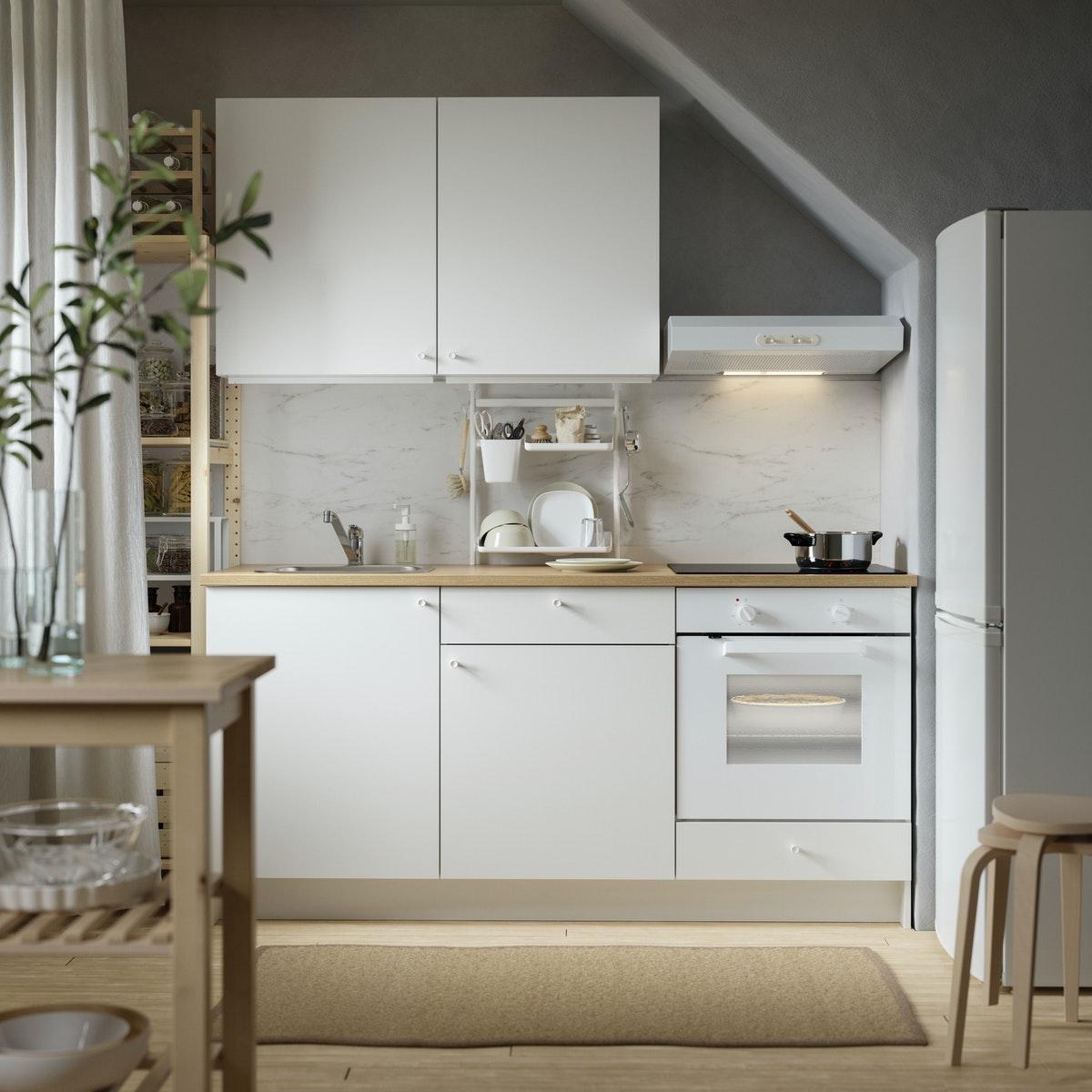 Zusehen ist eine kleinerer IKEA Küchenlösung mit weißen Fronten an den Unter- und Oberschränken mit einer Hölzernen Arbeitsplatte. Rechts davon ein IKEA Kühlschrank. Davor ein bescher Teppich. Im vorderen Teil des Bildes ist ein Holztisch mit einer Dekopflanze zusehen.