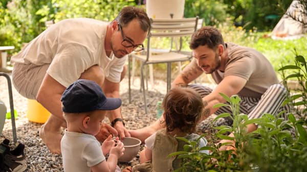 Zwei Erwachsene knien im Garten auf Kiesboden und spielen mit zwei Kleinkindern bei sommerlichem Wetter. Im Hintergrund stehen Gartenmöbel und viel Grün.