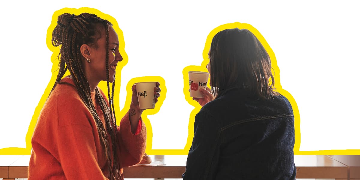 Zwei Frauen sitzen im Schwedischen Restaurant, lachen miteinander und trinken aus HEJ Papierbechern während einer entspannten Auszeit.