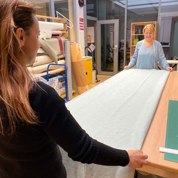 Zwei Frauen stehen an einem grossen Schneidertisch gegenüber und ziehen eine grosse, graue Stoffbahn glatt aus der ein Vorhang genäht werden soll