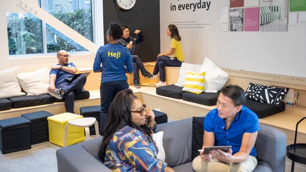 Zwei IKEA Mitarbeitende in Alltagskleidung sitzen auf einer großen hölzernen Treppe, trinken Kaffee und unterhalten sich.