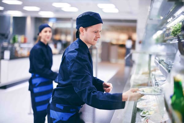 Zwei IKEA Mitarbeitende in dunkelblauen Uniformen aus dem Restaurant füllen die Salattheke auf. 