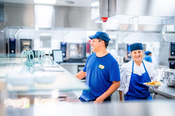 Zwei IKEA Mitarbeitende in dunkelblauen Uniformen lachen, während sie eine kleine Essenspause machen.