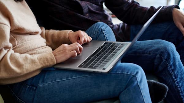 Zwei Personen sitzen auf einem Sofa, eine davn hat einen geöffneten Laptop auf dem Schoss