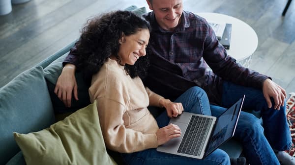 Zwei Personen sitzen lachend auf einem Sofa und schauen in den Bildschirm eines aufgeklappten Laptops den die eine Person auf dem Schoss stehen hat