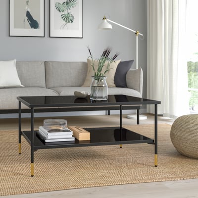 Modern living room with black & gold coffee table, glass top, surrounded by grey sofa, cushions, and natural light curtained window.