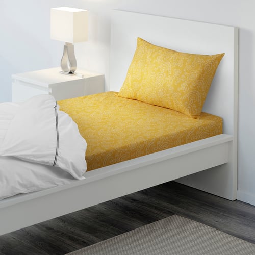 Bed with bright yellow patterned pillow, white sheet, and white bed frame. Bedside table and lamp beside it.
