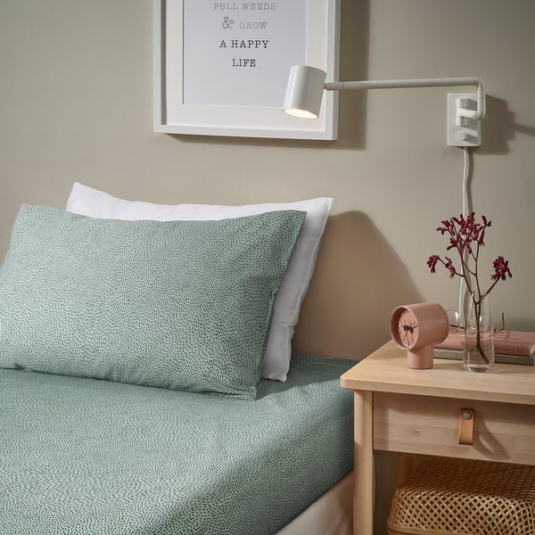 Cosy bedroom with ALPTALL green dot bedding, white wall lamp, pink clock, flowers in vase, and books on small wooden bedside table.