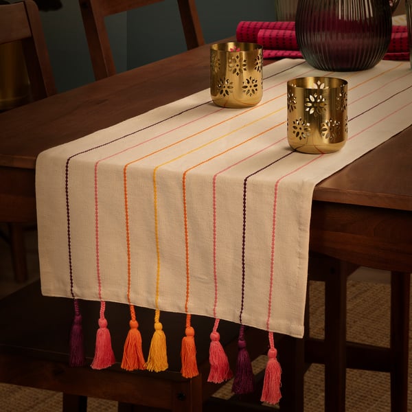 Beige table runner with colourful stripes and tassels. Gold decorative candle holders on top. Creates festive atmosphere.