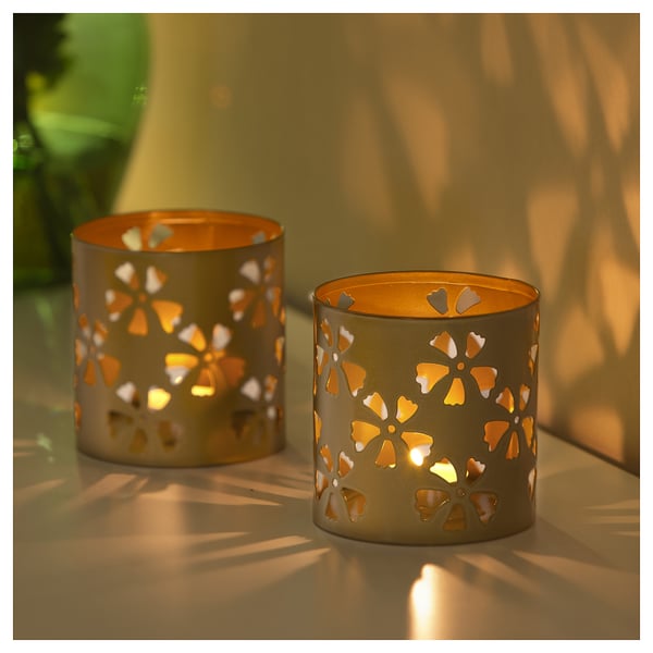 Two golden cylindrical tealight holders with cut-out flower designs cast decorative shadows when lit.