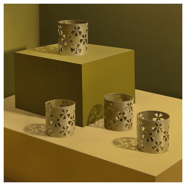 Gold-colored metal tealight holders with cut-out floral designs sit on a beige surface. Three holders of different sizes.