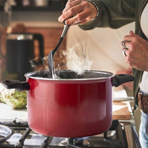 Person stirs boiling pot on stove with steam rising. Red pot has black handles, lid with holes.